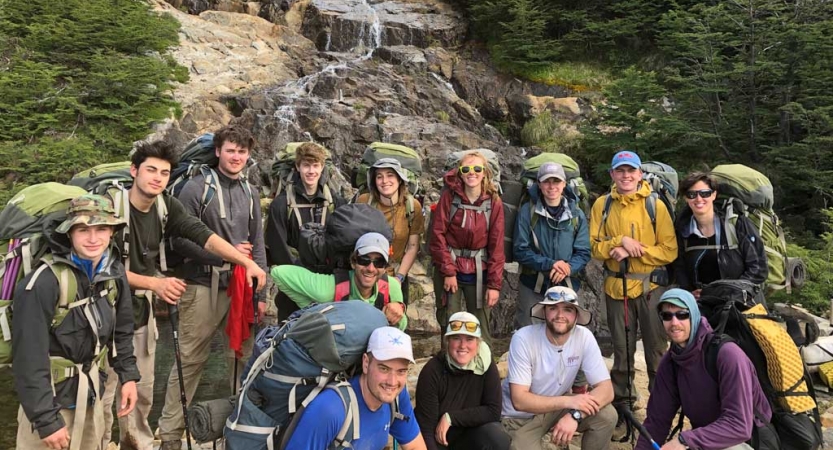 A group of people wearing backpacks pose for a photo in front of a small waterfall.